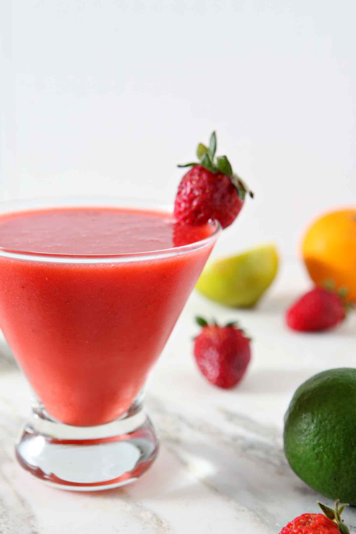 A Strawberry Virgin Margarita sits on a marble background, surrounded by strawberries, limes and oranges.