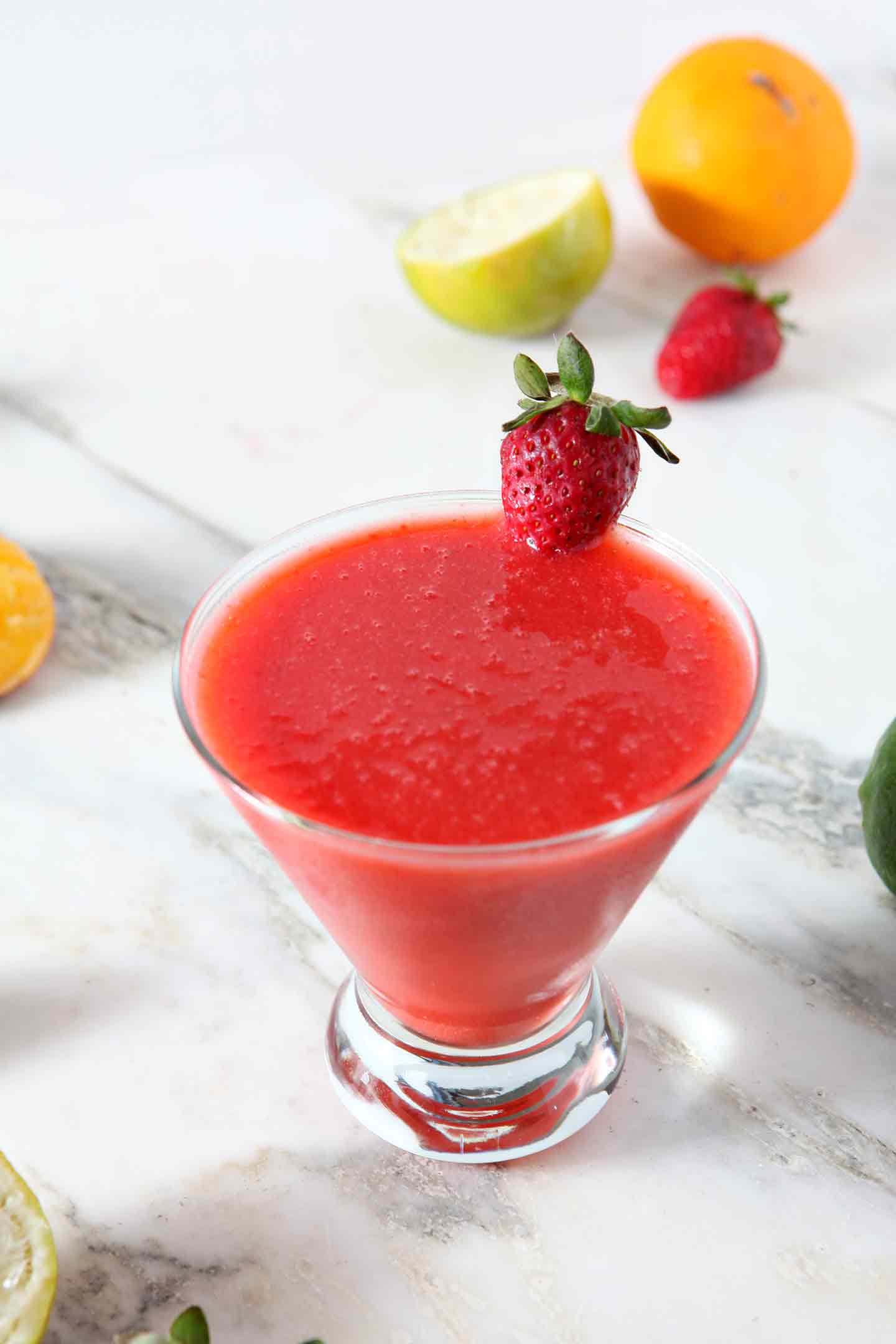 A Strawberry Virgin Margarita sits on a marble background, surrounded by strawberries, limes and oranges.