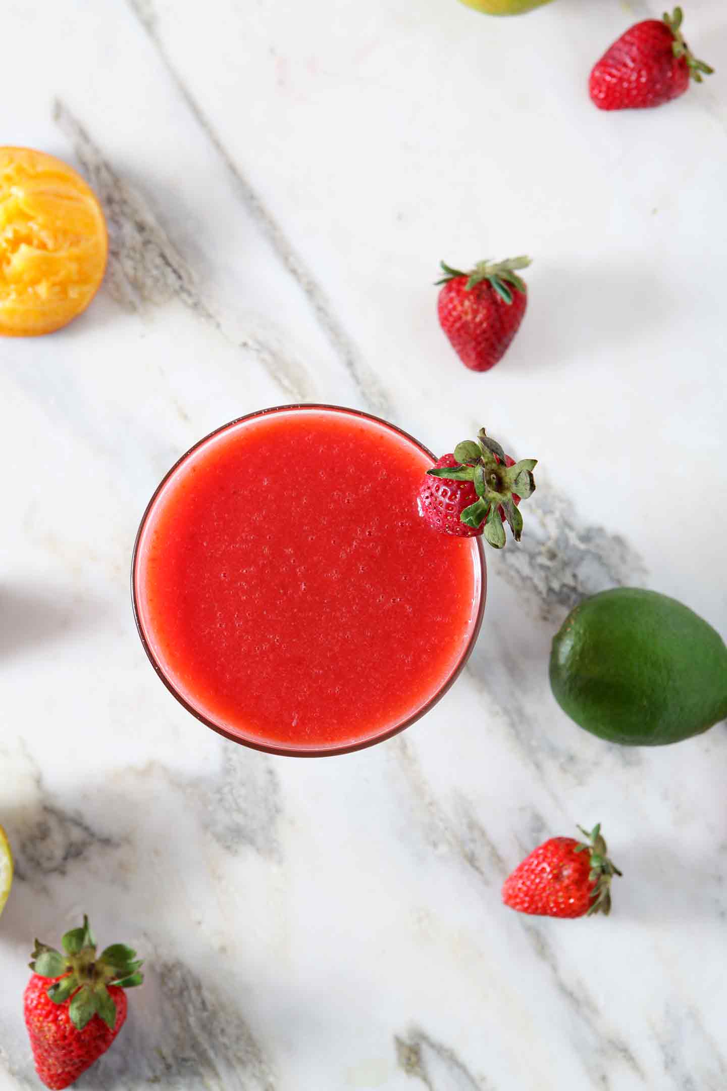 A Strawberry Virgin Margarita sits on a marble background, surrounded by strawberries, limes and oranges.