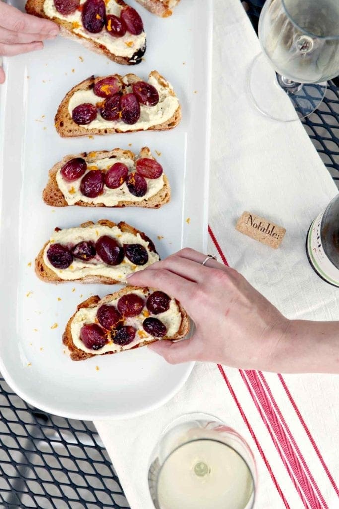 A person grabs a Roasted Grape Bruschetta from a white platter before consuming