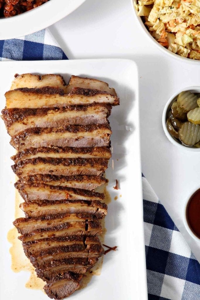 Overhead image of Instant Pot BBQ Brisket, served on a white platter, with pickles, barbecue sauce, coleslaw and baked beans