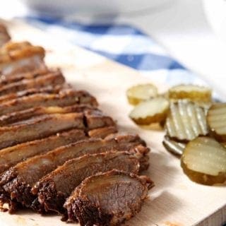 Sliced Instant Pot BBQ Brisket sits on a wooden cutting board with pickles and is served with baked beans, as shown in the background.