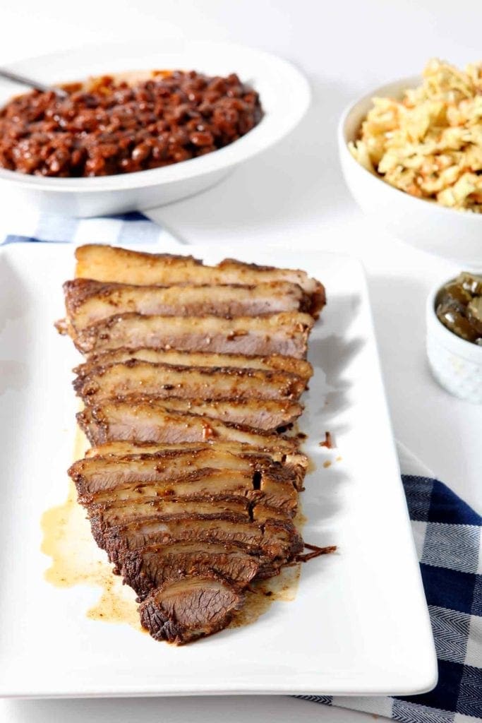 Instant Pot BBQ Brisket, served on a white platter, surrounded by barbecue sides like coleslaw, baked beans, pickles and barbecue sauce.