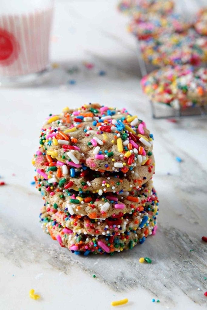 A stack of Funfetti Cookies are displayed on a marble background, shown with sprinkles, additional cookies and a glass of milk