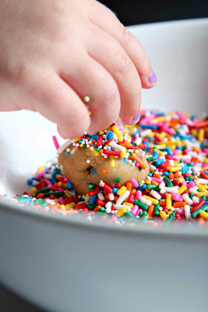 A small child rolls a ball of Funfetti Cookie Dough in additional sprinkles.