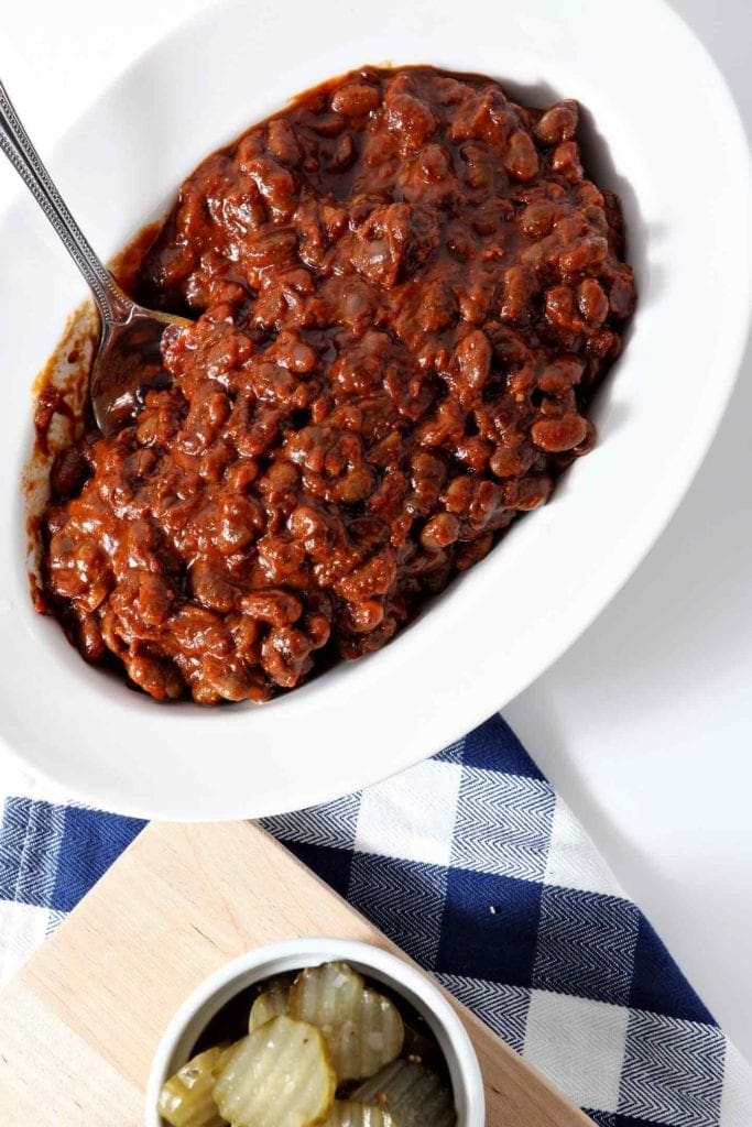 A bowl of BBQ Baked Beans, shown in a white bowl, shown with a blue buffalo plaid napkin and a platter of barbecue sides