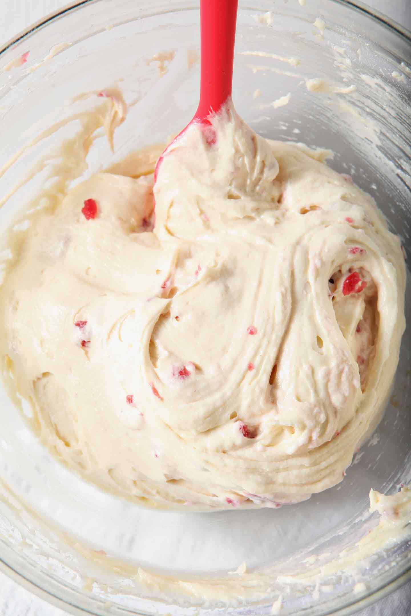 Overhead image of Mini White Chocolate-Cherry Bundt Cake (Instant Pot Cake) batter, shown in a clear glass bowl with a small red spatula