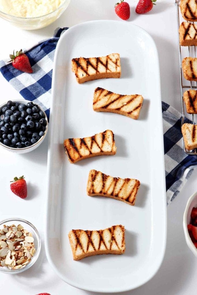 Grilled angel food cake, with apparent grill marks, sit on a white platter, surrounded by ingredients that will be used to make Patriotic Strawberry Bruschetta.