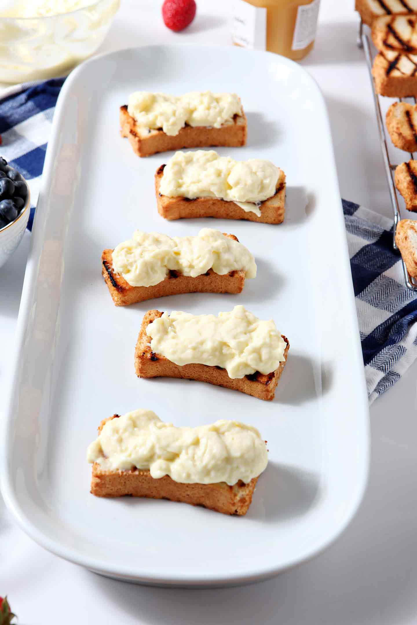 Pudding is carefully added to the toasted angel food cake slices for the Patriotic Strawberry Bruschetta, shown on a white platter, sitting on top of a blue buffalo print napkin