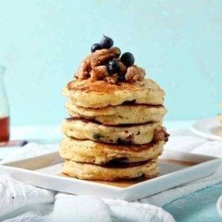 A stack of Blueberry Breakfast Sausage Pancakes sits on a square plate, covered in maple syrup.