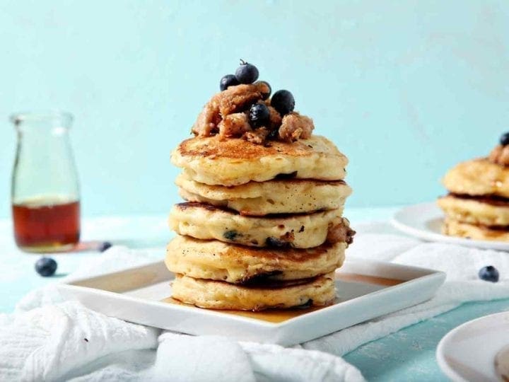 A stack of Blueberry Breakfast Sausage Pancakes sits on a square plate, covered in maple syrup.