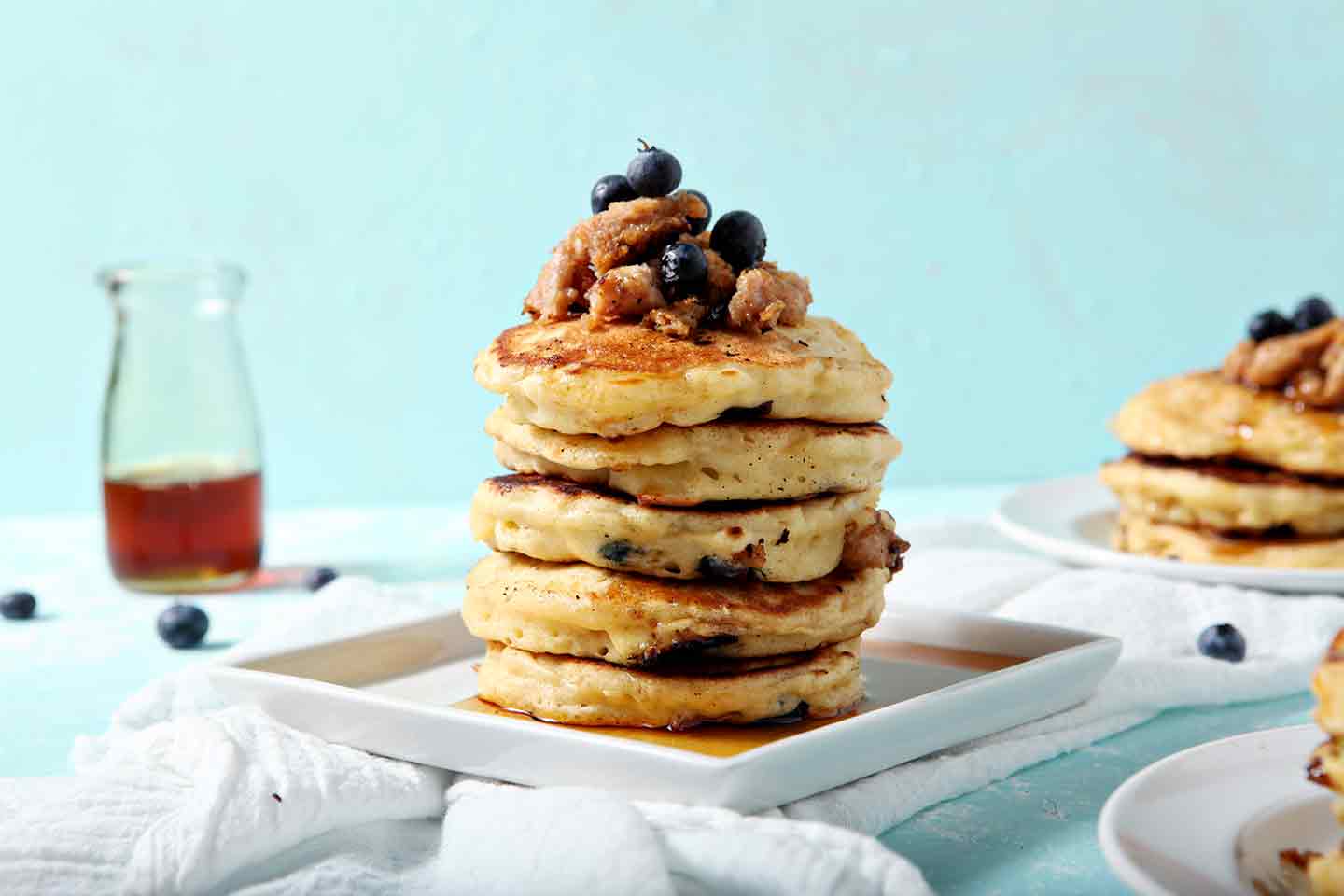 A stack of Blueberry Breakfast Sausage Pancakes sits on a square plate, covered in maple syrup.