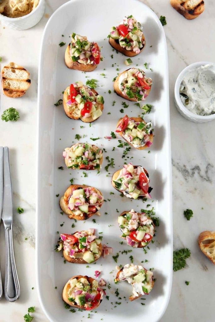 Overhead image of Greek Bruschetta, on a long white platter, surrounded with more bread slices, hummus and tzatziki.