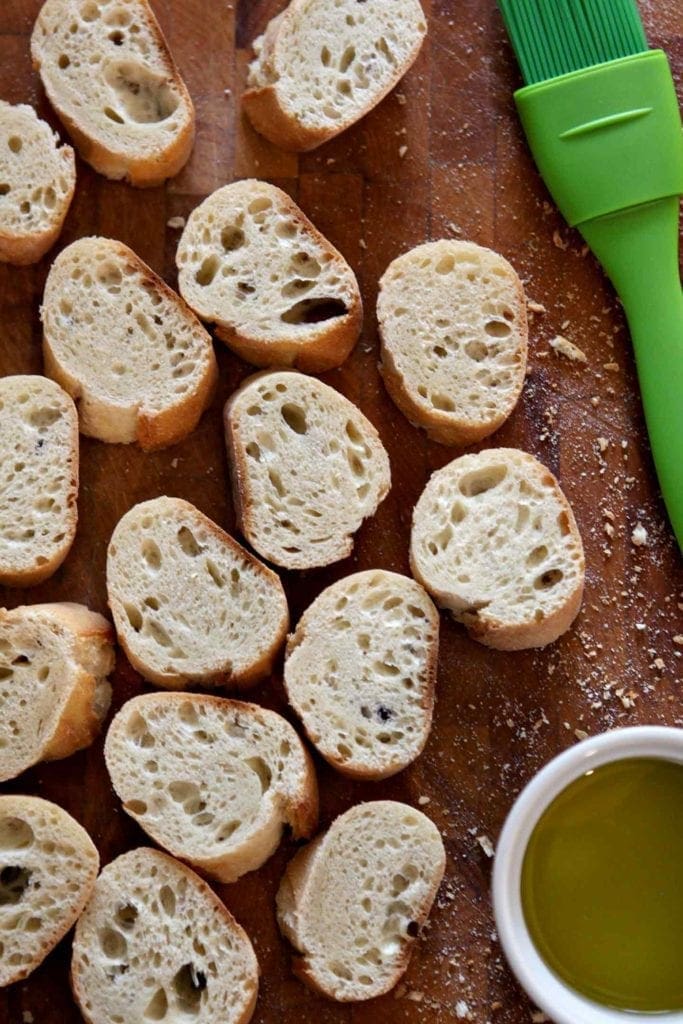 Bread slices sit on a cutting board with a bowl of olive oil and a pastry brush before grilling