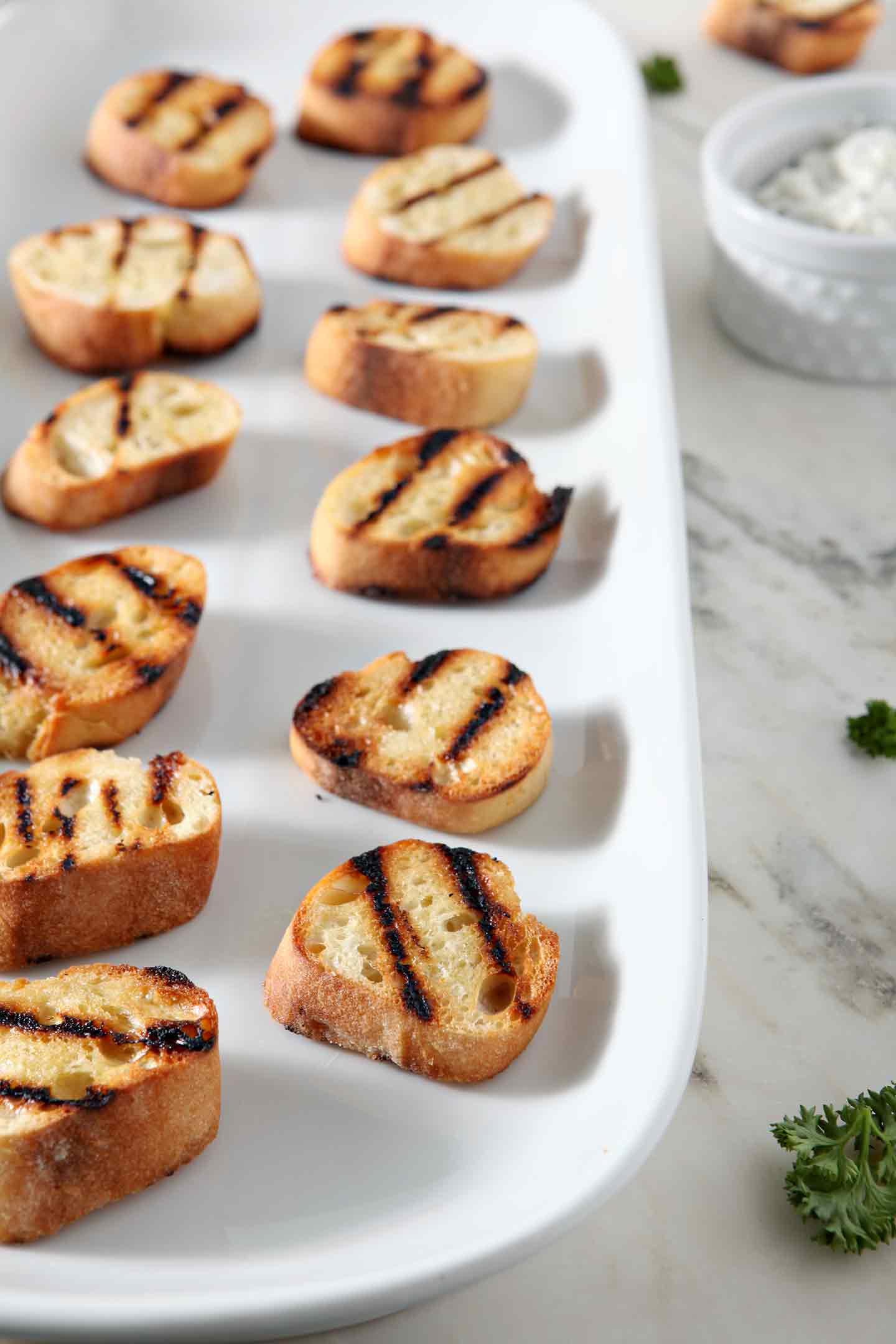 Grilled bruschetta rounds sit on a white porcelain platter before being topped with ingredients