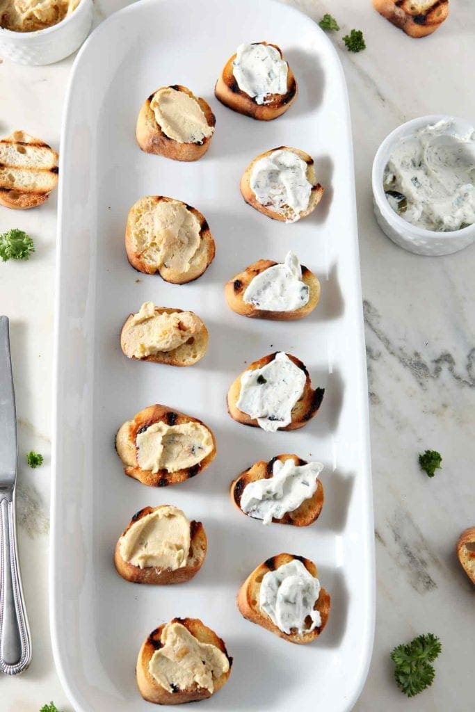 Hummus and tzatziki sauce are spread on top of toasty baguette rounds before bruschetta is assembled.