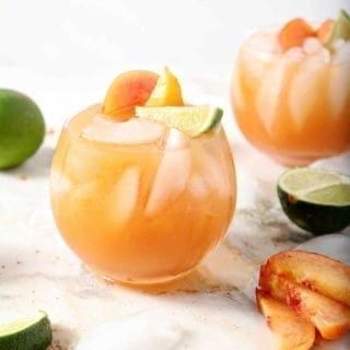 Two Peach Moscow Mule sit on a marble background, surrounded by peach slices and limes