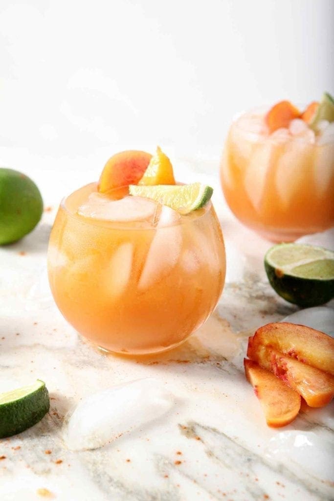 Two Peach Moscow Mule sit on a marble background, surrounded by peach slices and limes