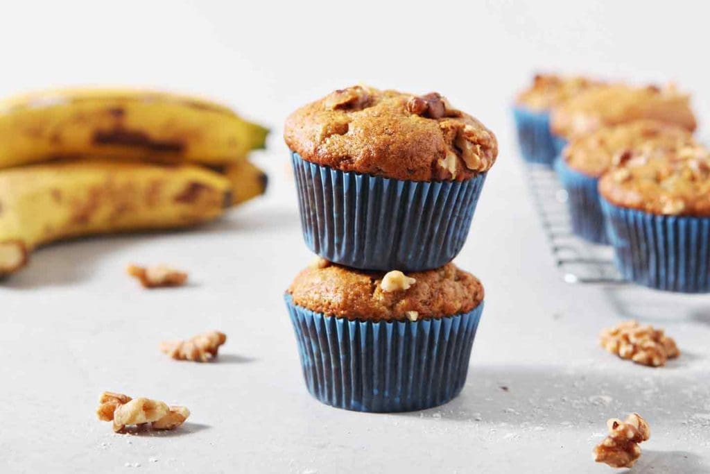 two muffins stacked on top of one another with bananas and other muffins in the background