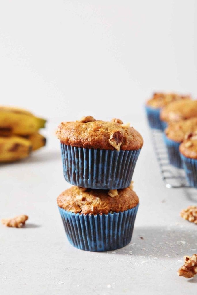 Two Vegan Banana Muffins are stacked on top of one another. Bananas and other muffins appear in the background.