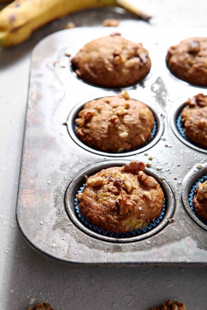 An entire muffin tin of Vegan Banana Muffins sits on a grey background, surrounded with bananas and walnuts, before serving