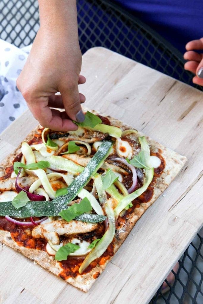 A woman adds fresh basil leaves to Grilled BBQ Chicken Flatbread before slicing it