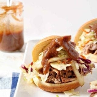 Three Pear BBQ Pulled Pork Sandwiches with Pear Slaw sit on a white platter with a BBQ sauce jar in the background