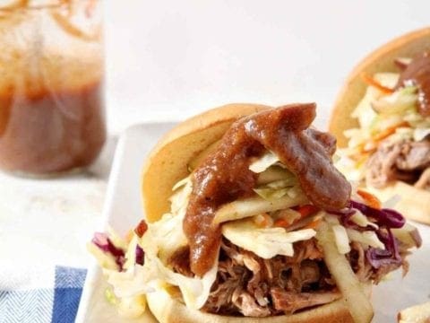 Three Pear BBQ Pulled Pork Sandwiches with Pear Slaw sit on a white platter with a BBQ sauce jar in the background