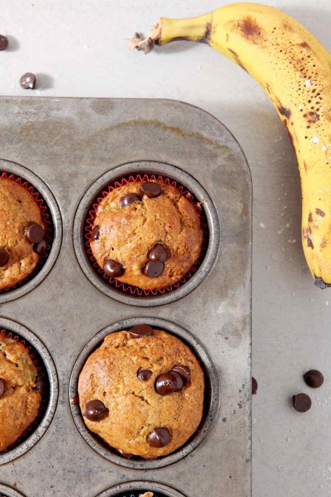 Vegan Banana Chocolate Chip Muffins, fresh from the oven, remain in the muffin tin, surrounded by chocolate chips and bananas