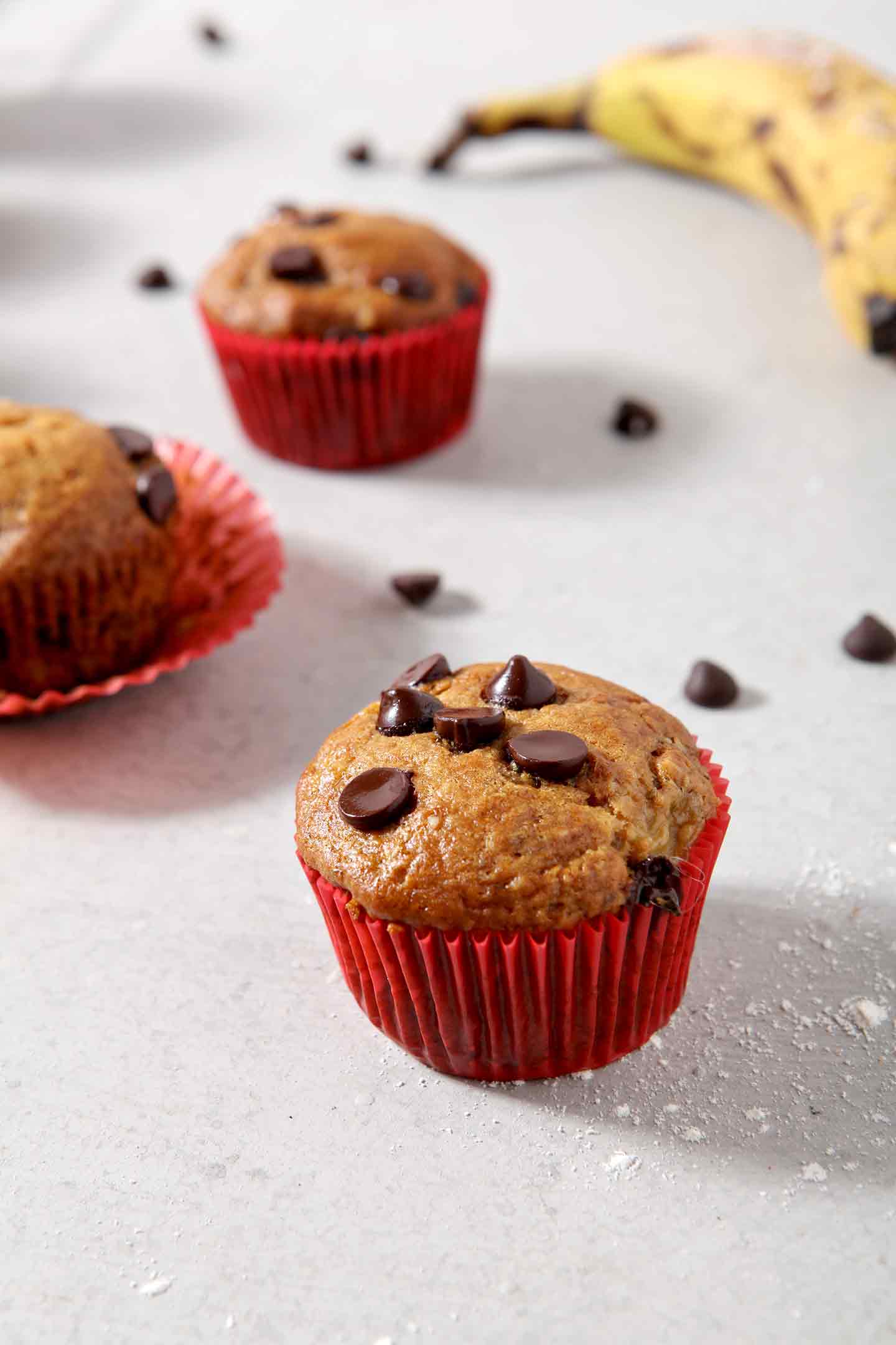 Three Vegan Banana Chocolate Chip Muffins sit on a grey background, surrounded by chocolate chips and bananas