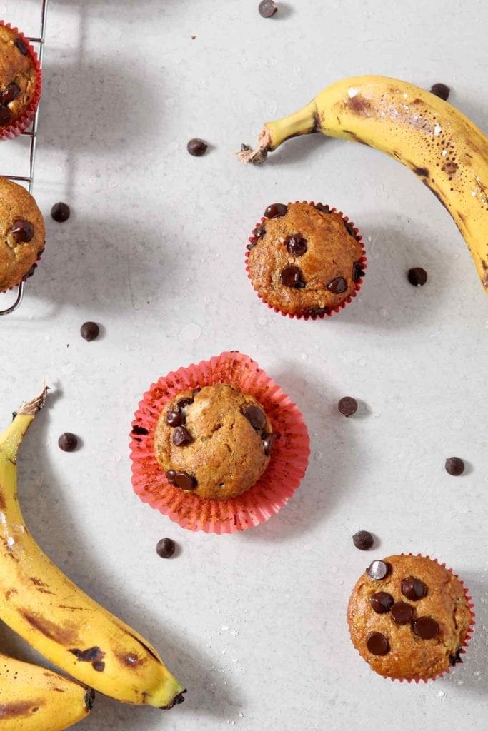 Overhead image of Vegan Banana Chocolate Chip Muffins, on a grey background, surrounded by chocolate chips and bananas