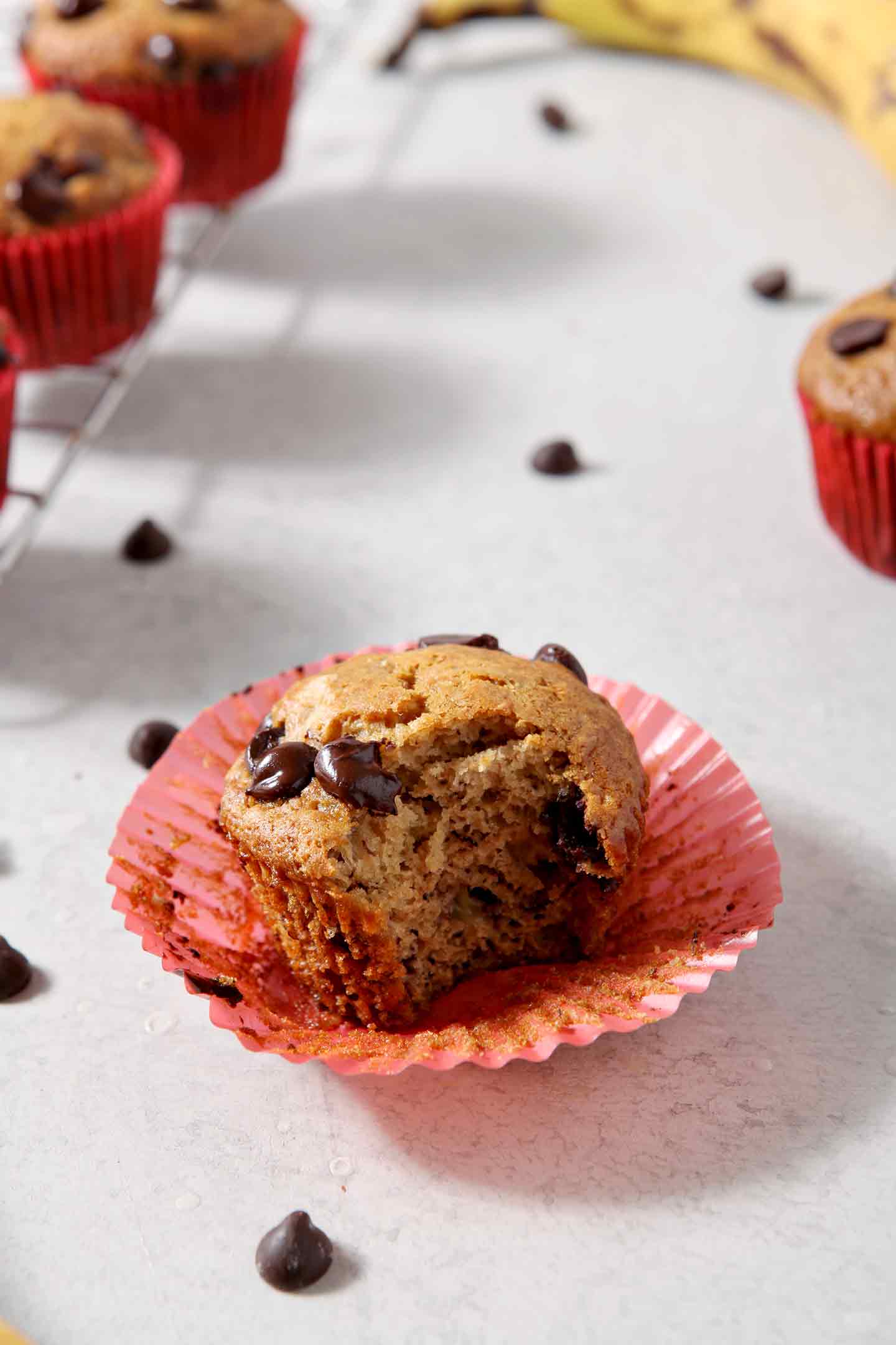 A bite mark is shown in a Vegan Banana Chocolate Chip Muffin, surrounded by other muffins, bananas and chocolate chips