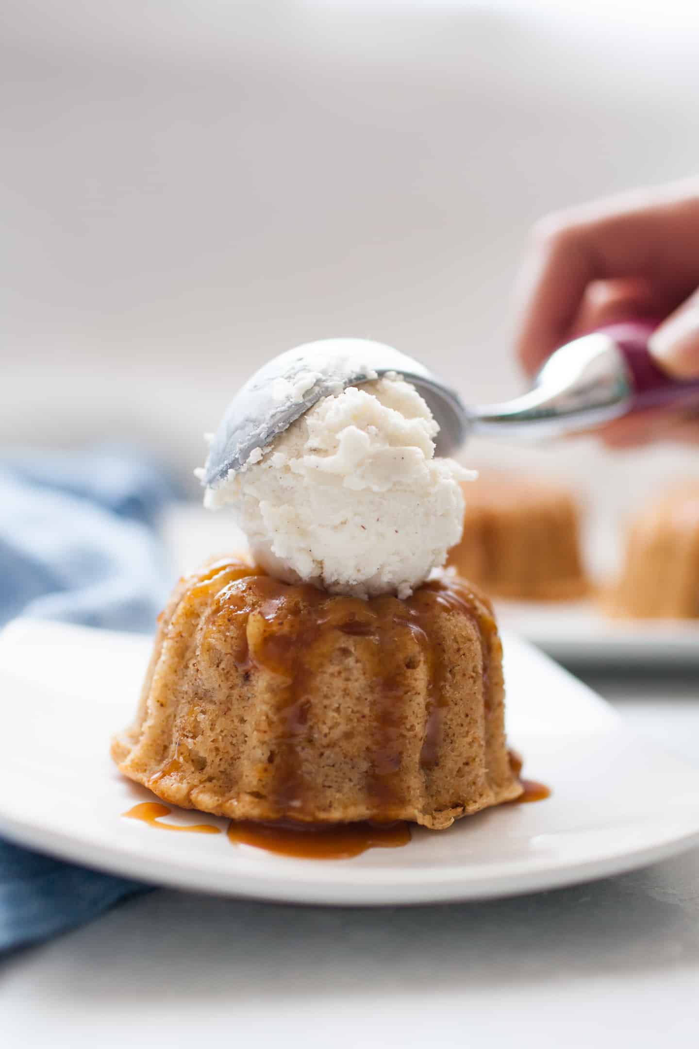 Scooping vanilla bean ice cream onto a mini spiced bundt cake sitting on a white square plate.