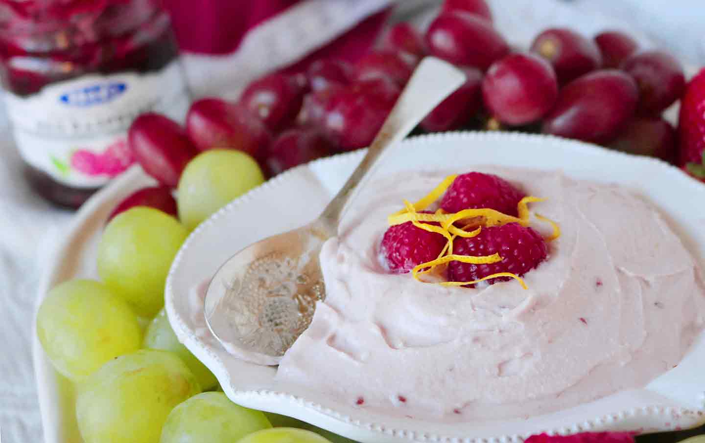 A bowl of Raspberry Yogurt Fruit Dip is served with a spoon and various fruits