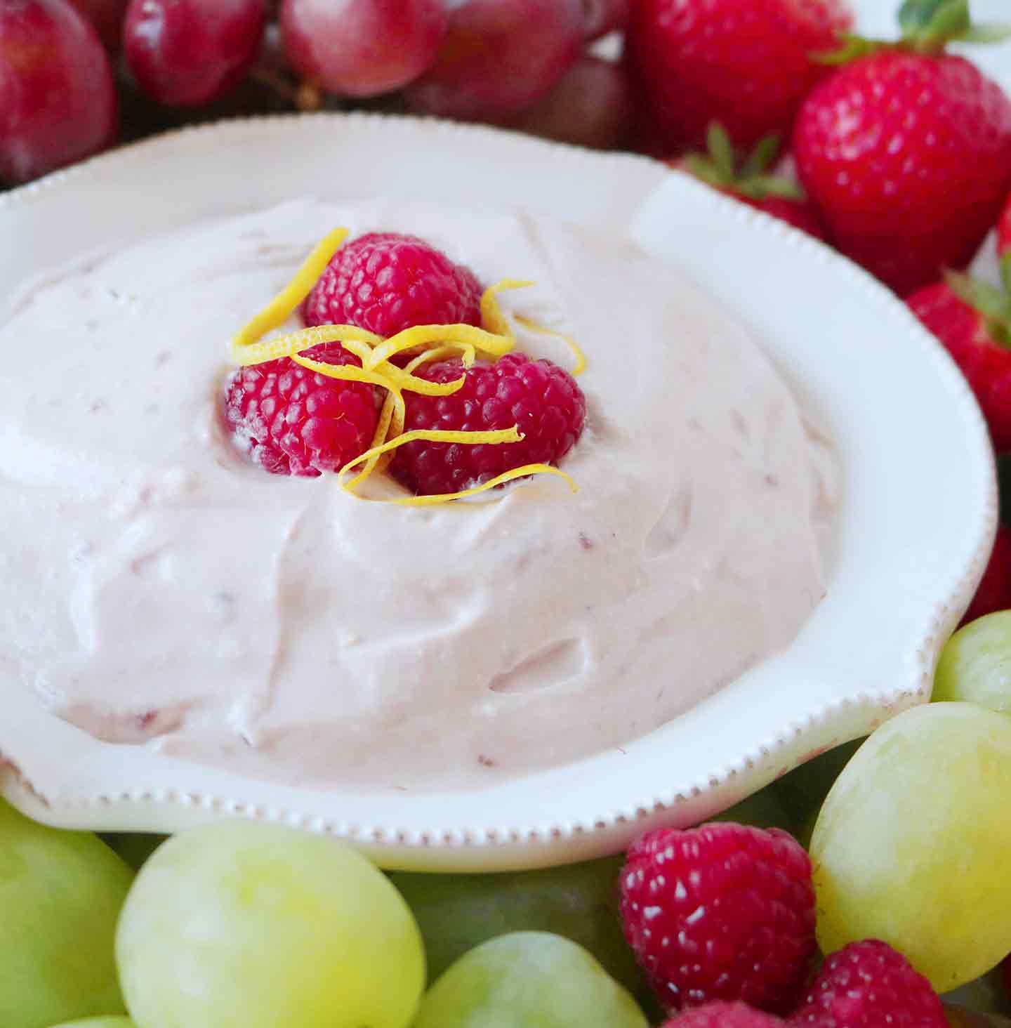 A bowl of Raspberry Yogurt Fruit Dip is served with green grapes, red grapes, fresh raspberries and strawberries