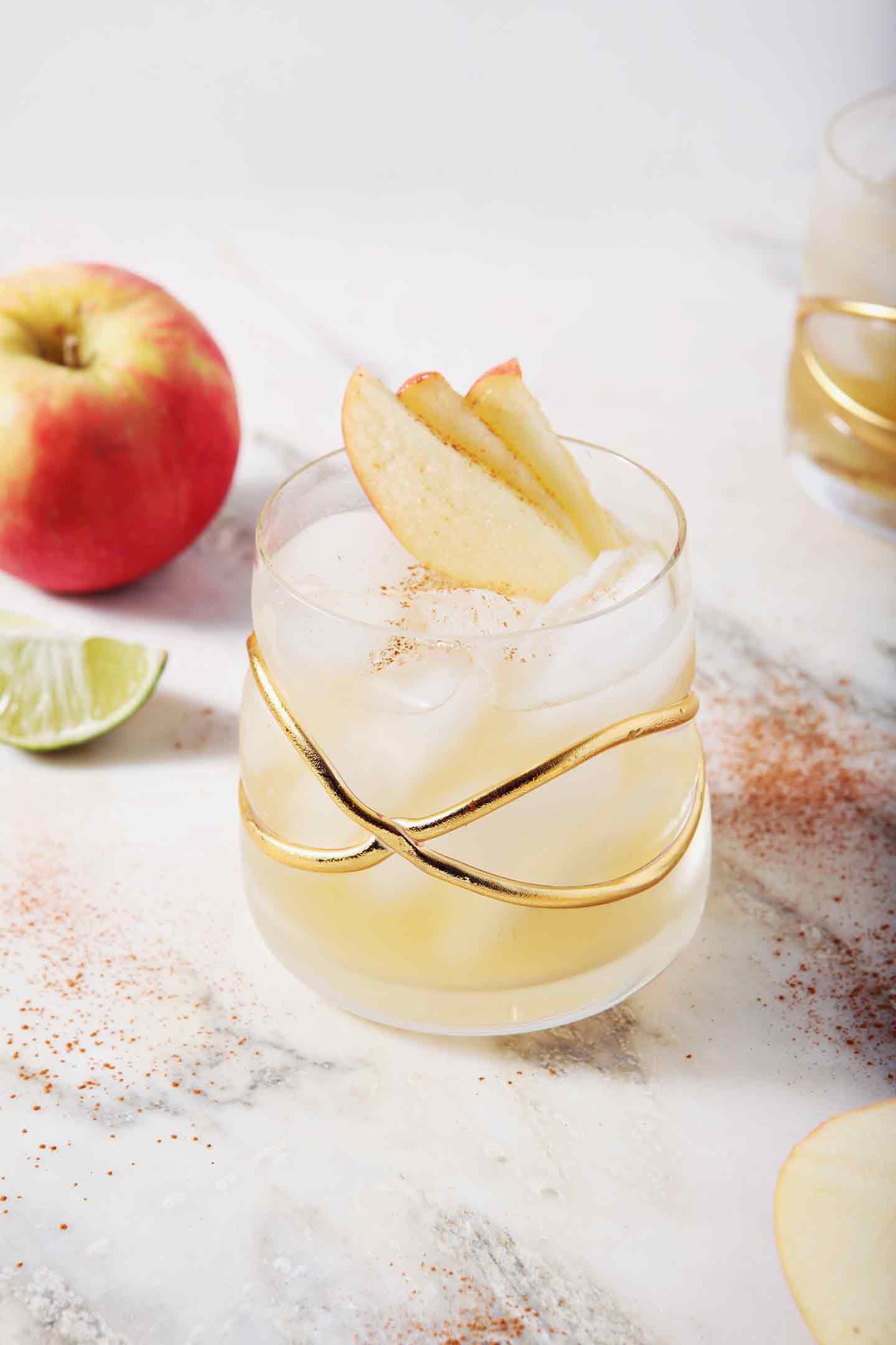 Two Apple Cider Moscow Mules sit on a marble background, surrounded by apples, lime slices and cinnamon