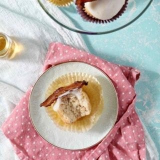 A bitten-into Bourbon Maple Bacon Cupcake lies on its side on a white plate, surrounded by more whole cupcakes