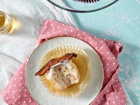 A bitten-into Bourbon Maple Bacon Cupcake lies on its side on a white plate, surrounded by more whole cupcakes