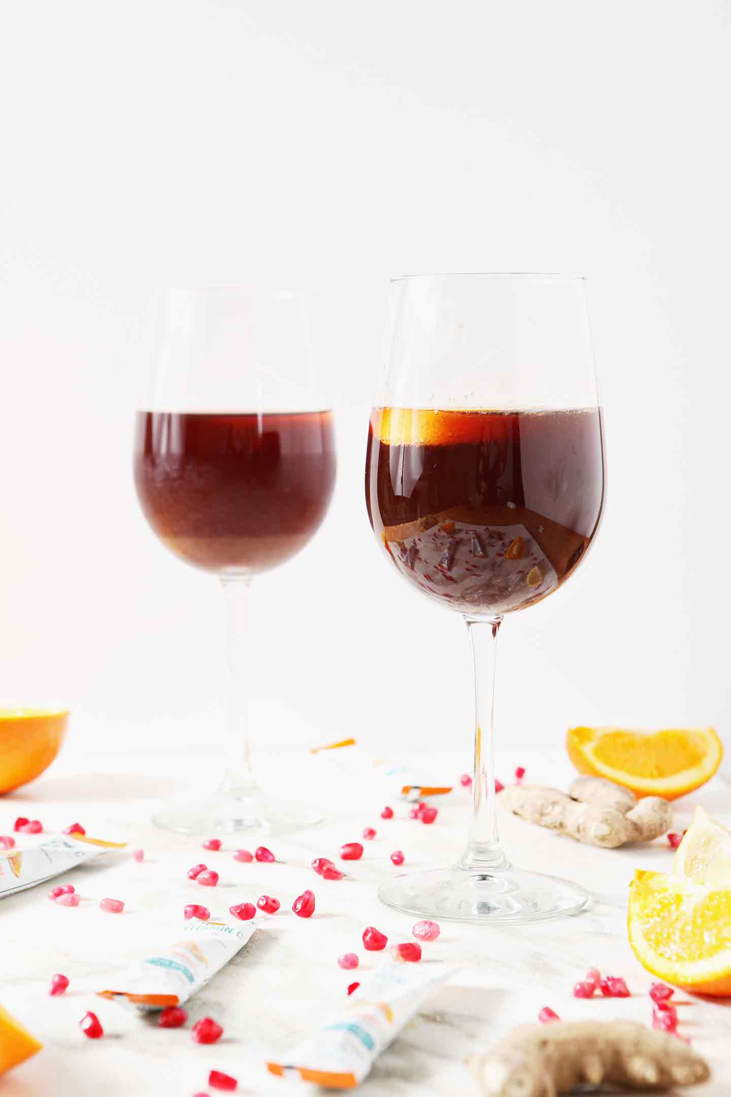 Two Pomegranate Ginger Orange Sparkling Mocktails sit on a marble background, surrounded by pomegranate arils and orange slices