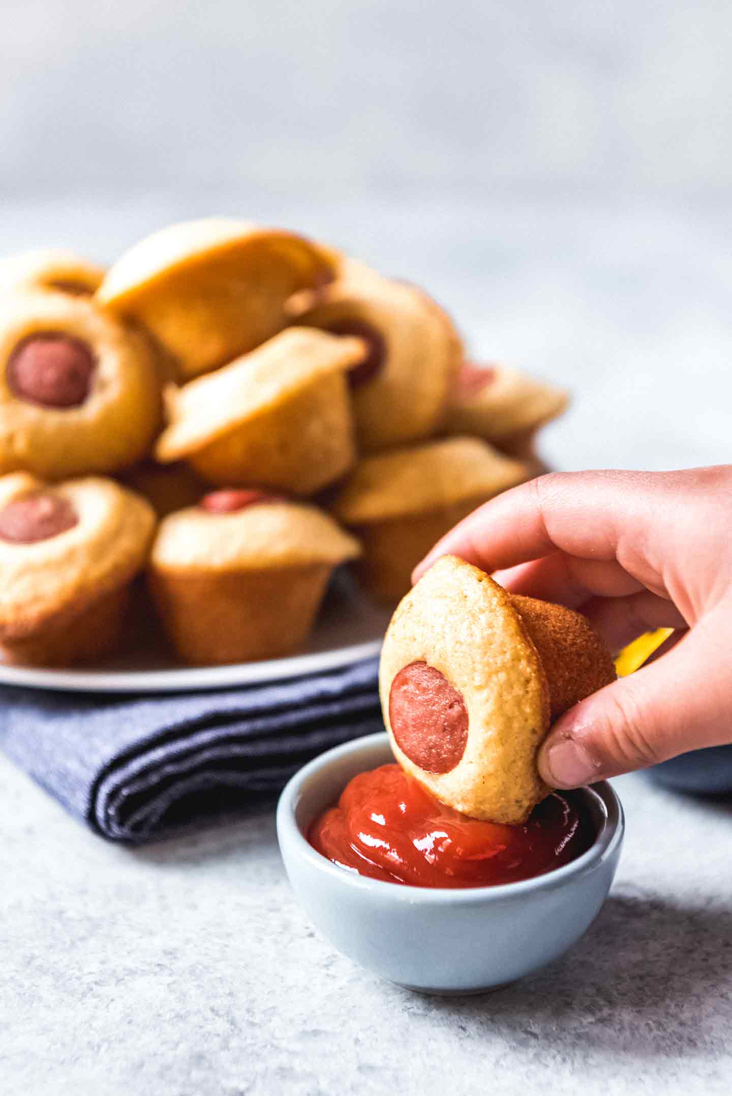 A person dips a Corn Dog Mini Muffin into ketchup with a plate of muffins shown behind it