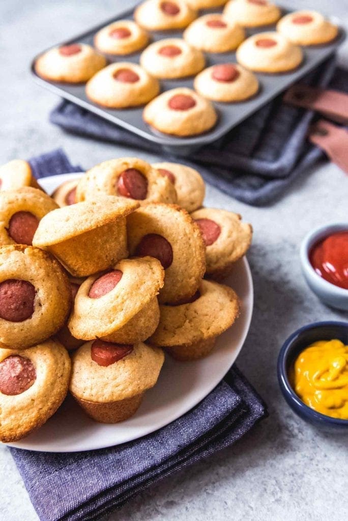 Corn Dog Mini Muffins are served on a plate with a muffin tin of them in the background, served with ketchup and mustard