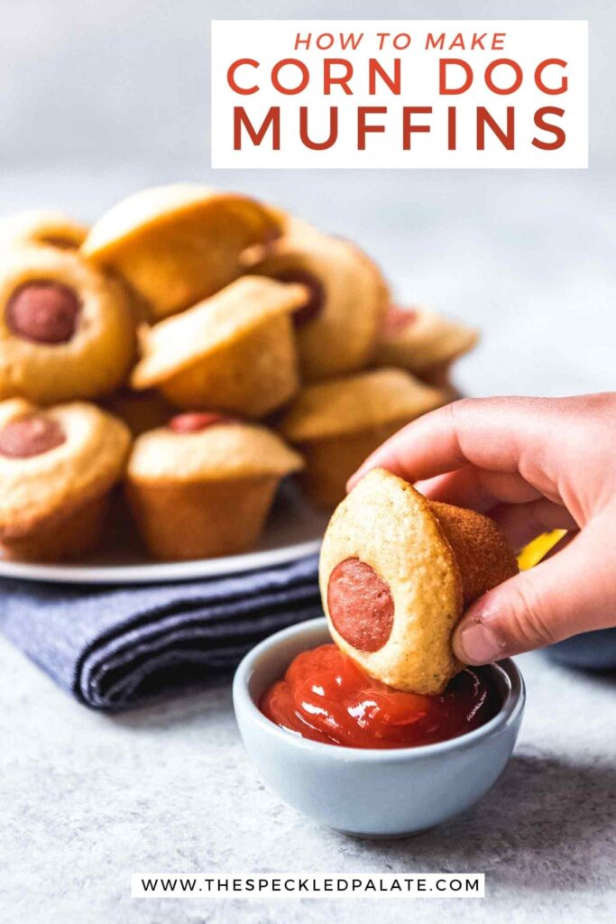 A Mini Corn Dog Muffin is dipped into a bowl of ketchup in front of a plate of more mini muffins with the text 'how to make corn dog muffins'