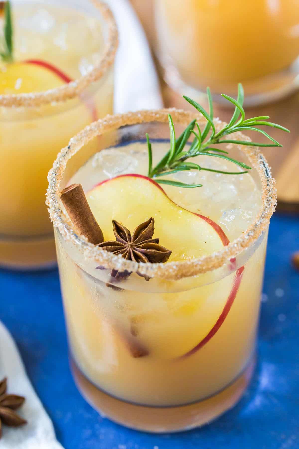 Close up of a Apple Cider Margarita on a blue background, garnished with an apple slice, cinnamon stick, star anise and sprig of rosemary