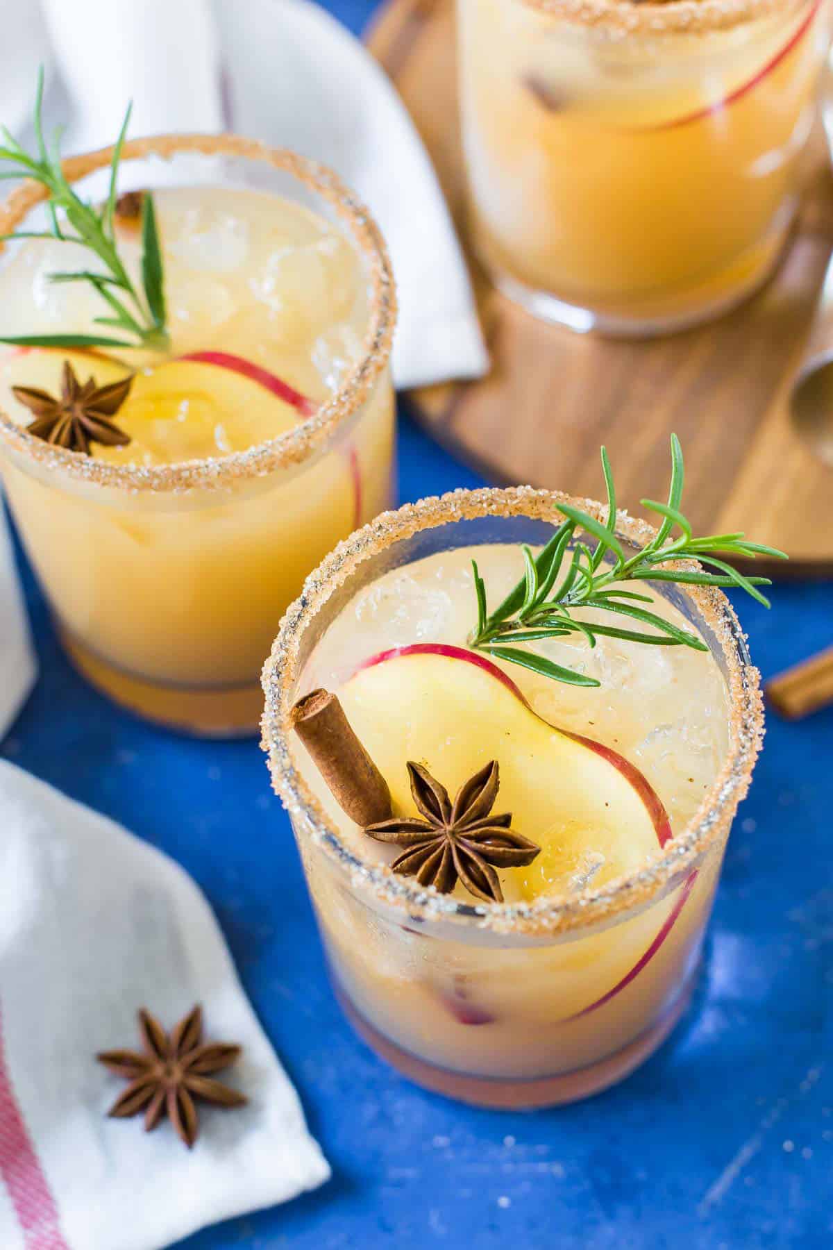 Close up of three Apple Margaritas on a blue background, garnished with an apple slice, cinnamon stick, star anise and sprig of rosemary