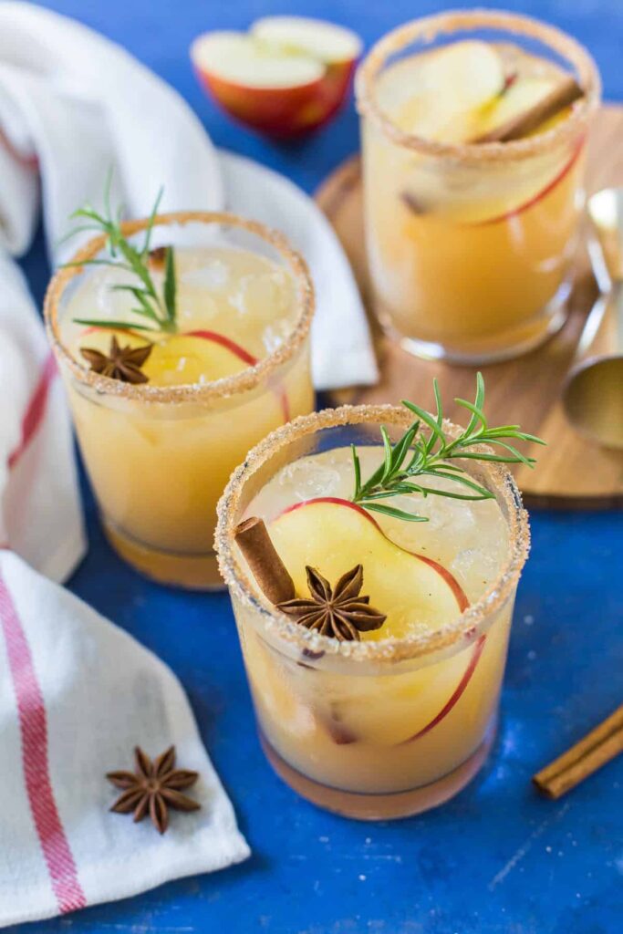 Three Apple Cider Margaritas, garnished with apple slices, cinnamon sticks, star anise and sprigs of rosemary, are served on a blue background