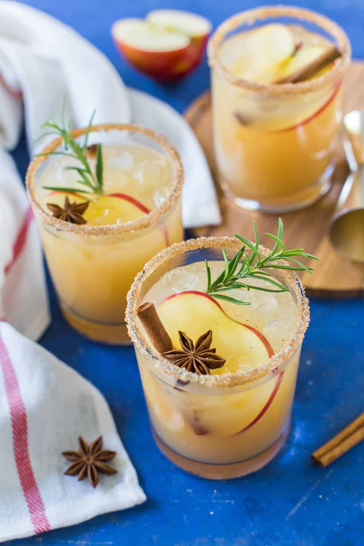 Three Apple Margaritas, garnished with apple slices, cinnamon sticks, star anise and sprigs of rosemary, are served on a blue background