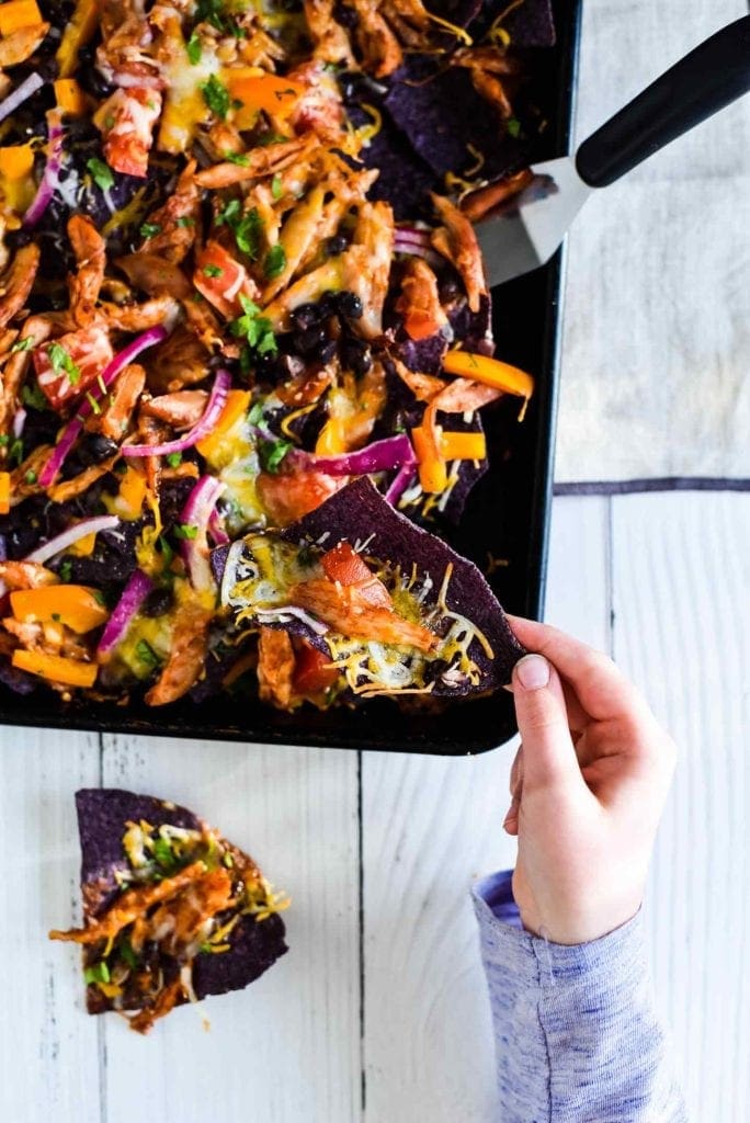 A girl grabs a chip off of a sheet pan holding Sheet Pan BBQ Chicken Nachos 