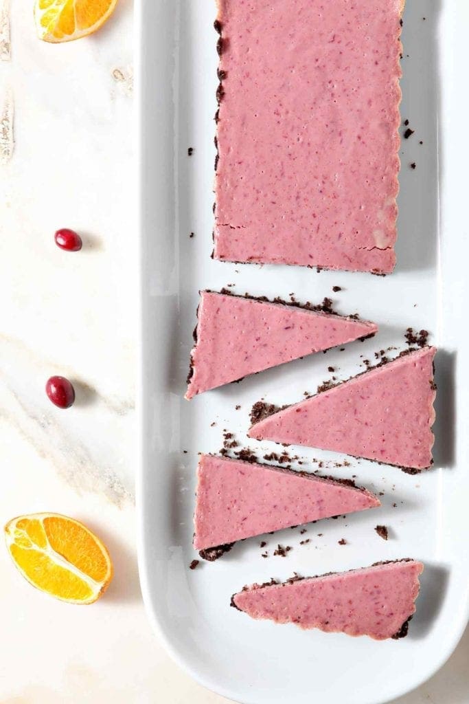 A partially sliced Creamy Cranberry Tart is served on a white platter, surrounded by orange slices and fresh cranberries