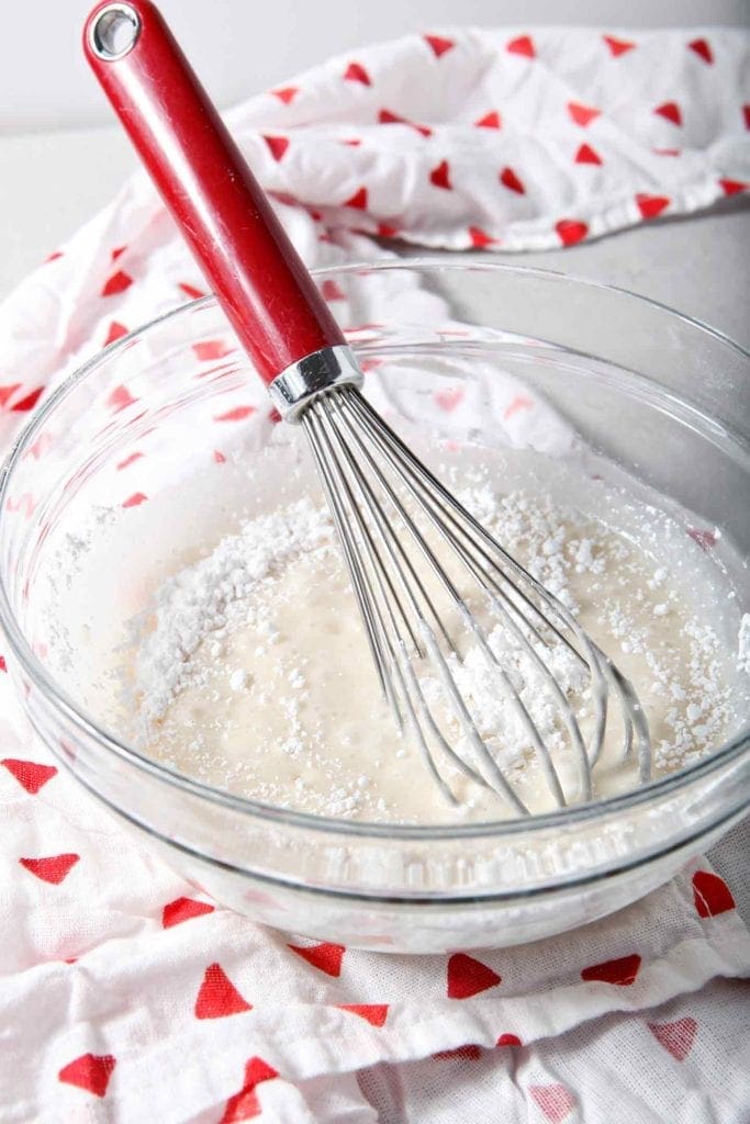 A red whisk is shown in a bowl as it mixes up Easy Sugar Cookie Icing