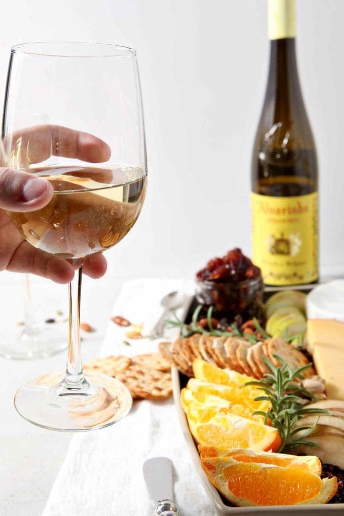 A person holds a glass of Vinho Verde wine in front of a Thanksgiving Cheese Board.
